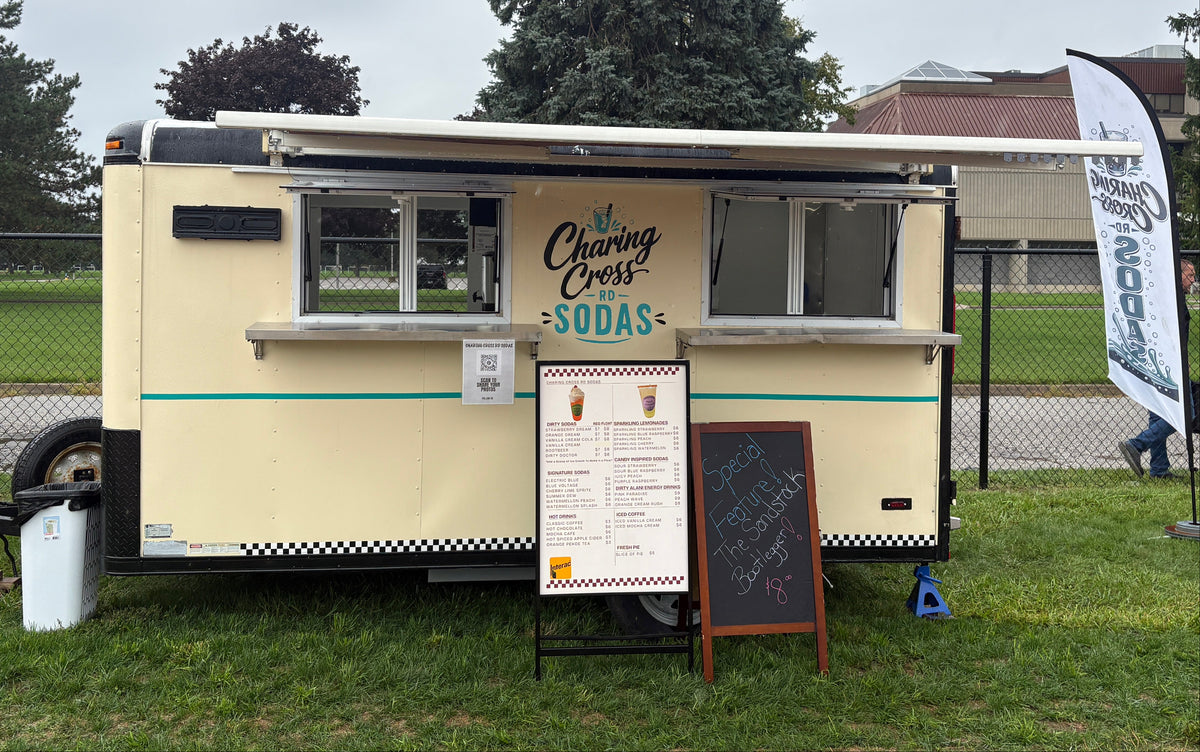 A vintage-style cream and black food trailer parked on grass outdoors, with a serving window, signage reading 'The Clearing Cafe - Sodas,' a menu board, chalkboard sign, and a tall white event banner on a cloudy day.